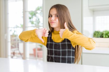 Genç güzel sarışın çocuk kız evde rahat sarı kazak giyen başparmak yukarı ve aşağı, anlaşmazlık ve anlaşma ifade yapıyor. Çılgın çatışma