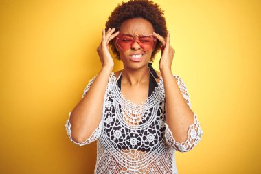 Afro saçlı genç afro-amerikalı kadın bikini ve kalp şeklinde güneş gözlüğü ağrı ve migren nedeniyle ağrı ve stresli baş ağrısı muzdarip. Eller baş üstünde.