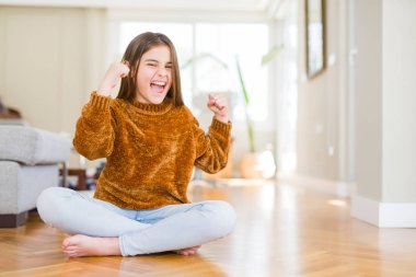 Güzel genç kız çocuk evde yerde oturan çok mutlu ve heyecanlı kolları ile kazanan jest yapıyor, gülümseyerek ve başarı için çığlık. Kutlama konsepti.