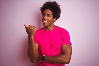 Afro saçlı, izole pembe arka plan üzerinde duran t-shirt giyen genç amerikalı adam mutlu yüz görünümlü gülümseyerek ve başparmak ile yan işaret.