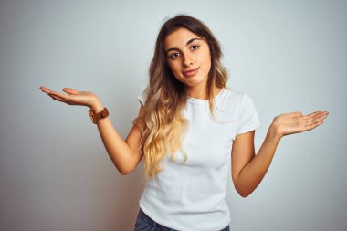 Genç güzel kadın izole arka plan üzerinde rahat beyaz t-shirt giyen habersiz ve kollar ve eller kaldırdı ile karışık ifade. Şüphe kavramı.