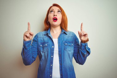 Beyaz izole arka plan üzerinde duran denim gömlek giyen genç güzel kızıl saçlı kadın şaşırttı ve yukarı bakarak ve parmakları ile işaret ve kolları kaldırdı şaşırttı.