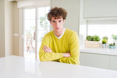 Evde sarı kazak giyen genç yakışıklı adam şüpheci ve sinirli, çapraz kolları ile yüzünde ki onaylamayan ifade. Negatif kişi.