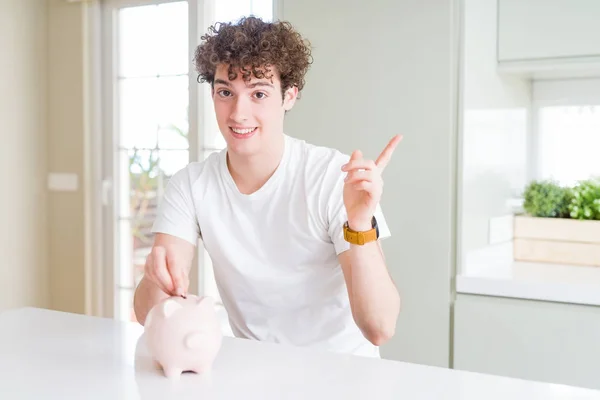 Young man investing money using piggy bank at home very happy pointing with hand and finger to the side