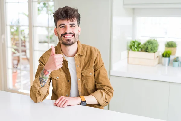 Young man wearing casual jacket sitting on white table doing happy thumbs up gesture with hand. Approving expression looking at the camera showing success.