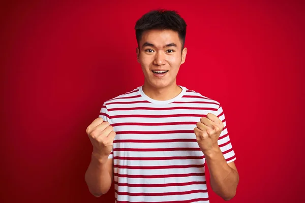 孤立した赤い背景の上に立つストライプのTシャツを着た若いアジアの中国人男性は 腕を上げて目を開けて成功に驚き 驚きました 勝者のコンセプト — ストック写真