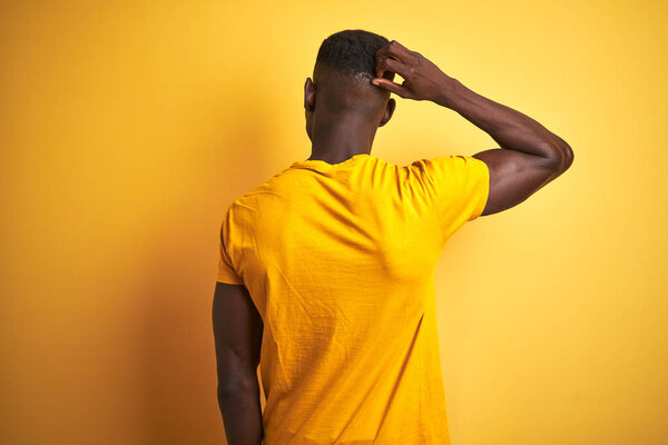 Young african american man wearing casual t-shirt standing over isolated yellow background Backwards thinking about doubt with hand on head