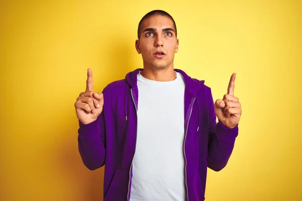 年轻的英俊男子穿着紫色运动衫在黄色孤立的背景惊讶和惊讶抬头 用手指指着和抬起手臂 — 图库照片