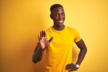 Genç afro-amerikan adam izole sarı arka plan waiving merhaba mutlu ve gülümseyen, samimi hoş geldin jesti söyleyerek üzerinde duran rahat t-shirt giyen