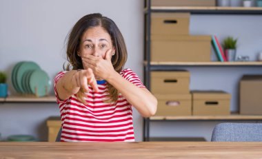 Orta yaş yaşlı kadın evde masada oturan size gülüyor, ağız üzerinde el ile kameraya parmak işaret, utanç ifade
