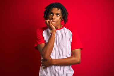 Çizgili t-shirt giyen dreadlocks ile Afro adam izole kırmızı arka plan üzerinde duran stresli ve ağız tırnak ısırma elleri ile sinir görünümlü. Anksiyete sorunu.