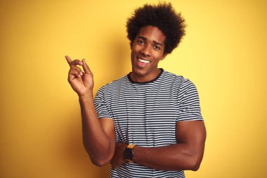 Afro saçlı Afro-Amerikan adam yüzünde büyük bir gülümseme ile izole sarı arka plan üzerinde lacivert çizgili t-shirt giyen, kameraya bakarak yan el ve parmak ile işaret.
