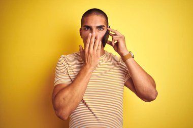 Genç yakışıklı adam sarı izole arka plan kapağı ağız üzerinde akıllı telefon kullanarak konuşurken el hata için utanç ile şok, korku ifadesi, sessizlik, gizli kavram korkmuş