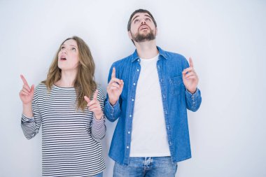 Genç güzel çift beyaz izole arka plan üzerinde birlikte ayakta şaşırmış ve baktı ve parmakları ile işaret ve kolları kaldırdı şaşırttı.