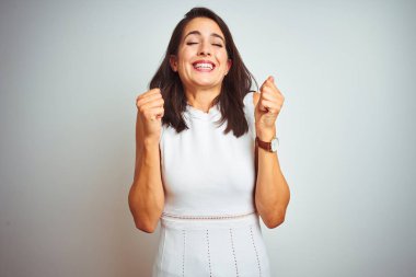 Beyaz izole arka plan üzerinde duran elbise giyen genç güzel kadın kollarını kaldırdı ve gözleri gülümseyerek zafer kutlayan kapalı başarı için heyecanlı. Kazanan kavramı.