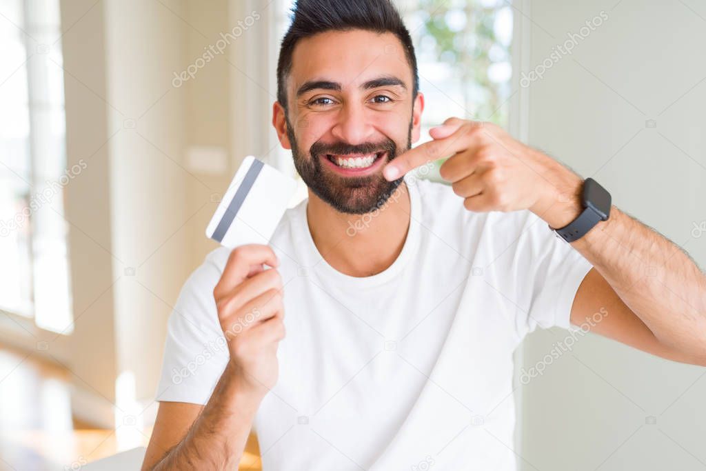 Handsome hispanic man holding credit card very happy pointing with hand and finger