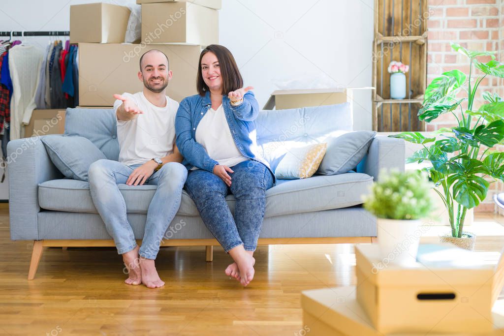 Young couple sitting on the sofa arround cardboard boxes moving to a new house smiling friendly offering handshake as greeting and welcoming. Successful business.