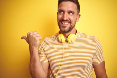 Genç yakışıklı adam sarı izole arka plan üzerinde müzik dinlerken kulaklık takan mutlu yüz görünümlü ve başparmak ile yan işaret gülümseyerek.