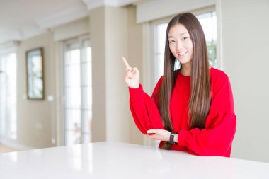 Yüzünde büyük bir gülümseme ile beyaz masada kırmızı kazak giyen güzel Asyalı kadın, kameraya bakarak yan el ve parmak ile işaret.