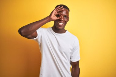 Beyaz tişört giyen genç afro-amerikan adam izole sarı arka plan üzerinde duran el gülümseyerek ok jest yapıyor, mutlu yüz ile parmakları ile bakarak göz.