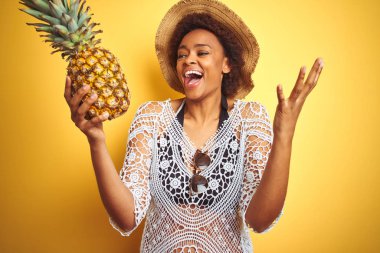 Sarı izole arka plan üzerinde pineaple tutan afro saçlı genç afro-amerikan kadın çok mutlu ve heyecanlı, kazanan ifade büyük bir gülümseme ile çığlık zafer kutluyor ve ellerini kaldırdı