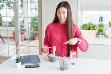 Güzel Asyalı kadın dizüstü bilgisayar kullanarak çalışan ve Asya noodle yeme çok mutlu el ve parmak ile işaret