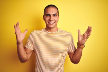 Genç yakışıklı adam sarı izole arka plan üzerinde çizgili t-shirt giyen deli kutluyor ve kollarını kaldırdı ve açık gözleri heyecan çığlık ile başarı için hayran. Kazanan kavramı