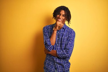 Izole sarı arka plan üzerinde duran rahat gömlek giyen dreadlocks ile Afro adam kamera çapraz kollar ve el çene üzerinde kaldırdı gülümseyerek emin görünümlü. Olumlu düşünme.