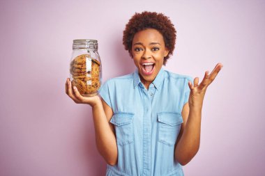 Pembe izole arka plan üzerinde çikolata cipsi kurabiye kavanoz tutan Afrikalı Amerikalı kadın çok mutlu ve heyecanlı, kazanan ifade büyük bir gülümseme ile çığlık zafer kutluyor ve ellerini kaldırdı