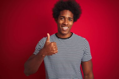 Afro saçlı genç Amerikalı adam izole kırmızı arka plan üzerinde lacivert çizgili t-shirt giyen el ile jest kadar mutlu başparmak yapıyor. Başarı göstererek kameraya bakan ifadeyi onaylama.