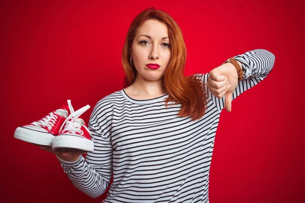 Joven Hermosa Pelirroja Sosteniendo Zapatillas Deporte Sobre Fondo Rojo Aislado —  Fotos de Stock