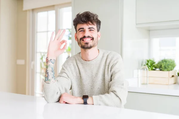 Young man wearing casual sweater sitting on white table smiling positive doing ok sign with hand and fingers. Successful expression.