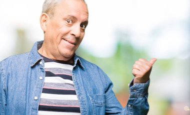 Yakışıklı kıdemli adam kot ceket giymiş mutlu yüz görünümlü ve başparmak ile yan işaret ile gülümseyen.