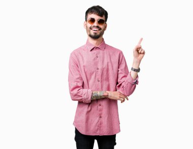 Kameraya bakarak yan el ve parmak ile işaret eden genç yakışıklı adam yüzünde kocaman bir gülümseme ile izole arka plan üzerinde güneş gözlüğü takıyor.