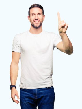 Yakışıklı adam giyiyor rahat beyaz t-shirt gösterme ve kendinden emin ve mutlu gülümseyen bir parmak ile işaret eden oktan yukarı.