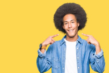 Afrikalı-Amerikalı genç afro saçlı emin gösterilen ve işaret parmakları diş ve ağız ile gülümseyen. Sağlık kavramı.
