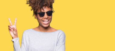 Güzel genç Afro-Amerikan kadın mutlu yüz zafer işareti yaparken kameraya göz kırpıyor ile gülümseyen izole arka plan üzerinde güneş gözlüğü takıyor. İki numaralı.