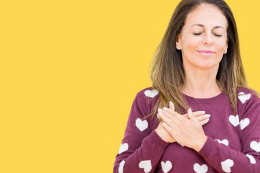 Göğüs kapalı gözlerle ellerinde ve yüzünde minnettar jest ile gülümseyen izole arka plan üzerinde kalpler kazak giyen güzel orta yaş kadın. Sağlık kavramı.