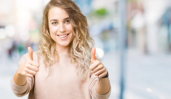 Beautiful young blonde woman wearing sweatershirt over isolated background approving doing positive gesture with hand, thumbs up smiling and happy for success. Looking at the camera, winner gesture.