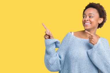 Gülümseyen ve iki elleri ve parmakları ile tarafına işaret kamera bakarak izole arka plan üzerinde bir kazak giyen genç güzel Afrikalı-Amerikalı kadın.