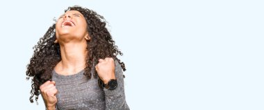 Kıvırcık saçlı genç güzel kadın gri kazak giyen çok mutlu ve heyecanlı kolları kaldırdı ile kazanan jest yapıyor, gülümseyerek ve başarı için çığlık. Kutlama konsepti.