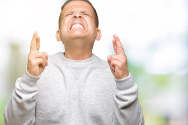 Orta yaş Arap geçiş parmakları umut ve gözleri kapalı ile gülümseyen izole arka plan üzerinde spor kazak giyen adam. Şans ve batıl kavramı.