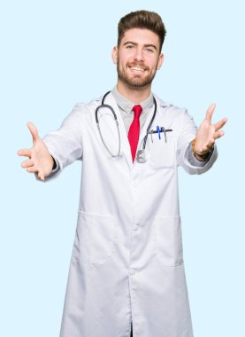 Genç yakışıklı sarılmak için kollarını açarak gülümseyerek kameraya bakarak tıbbi ceket giyen adam doktor. Neşeli ifade kucaklayan mutluluk.