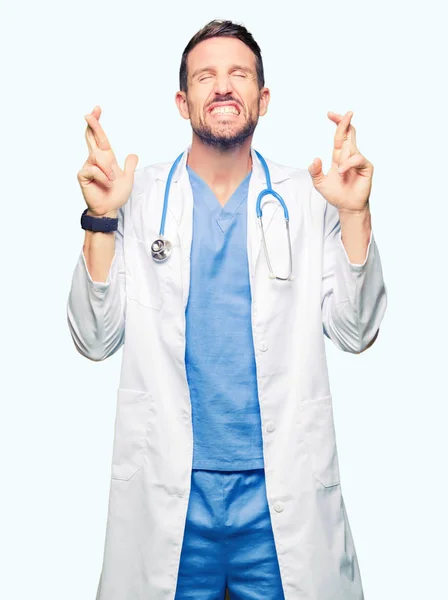 英俊的医生穿医疗制服在孤立的背景微笑交叉手指与希望和眼睛闭合 运气和迷信的概念 — 图库照片
