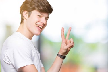 Mutlu yüz zafer işareti yaparken kameraya göz kırpıyor ile gülümseyen izole arka plan üzerinde rahat beyaz t-shirt giyen genç yakışıklı adam. İki numaralı.