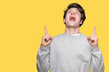 İzole arka plan üzerinde sweatshirt giyen genç yakışıklı sportif adam hayret ve yukarıya bakıyor ve parmakları ile işaret ve silah kaldırdı şaşırttı.