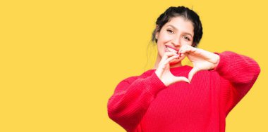 Kırmızı kazak ve kalp simgesi ve elleriyle şekil gösterilen aşık gülümseyen topuz giyen genç güzel kadın. Romantik kavramı.