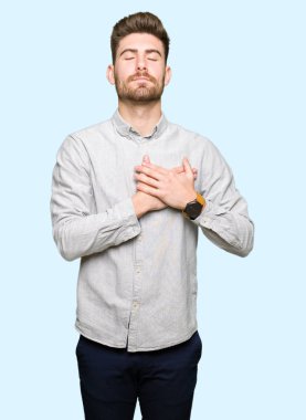 Gündelik tişört giyen, elleri göğsünde, gözleri kapalı gülümseyen ve yüzünde minnet ifadesi olan genç yakışıklı bir adam. sağlık kavramı.