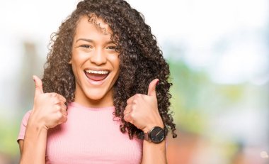 Pembe t-shirt başarı işareti giyen kıvırcık saçlı Genç güzel kadın el ile olumlu jest yapıyor, gülümseyen ve mutlu kadar başparmak. Neşeli ifade ile kamera bakarak, kazanan jest.