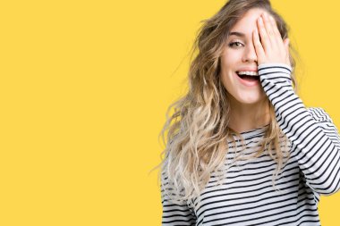 El yüz ve sürpriz duygu kendinden emin gülümseme ile tek gözüyle kapsayan izole arka plan üzerinde çizgili süveter giyen güzel genç sarışın kadın.